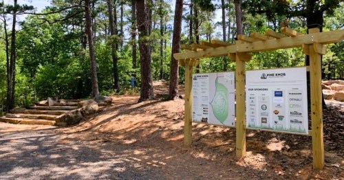 Signage at a wooded park entrance, featuring a map and sponsor logos, surrounded by trees and gravel paths.