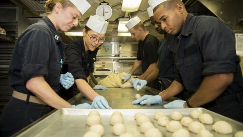 A group of people in chef hats and gloves prepare dough on a kitchen counter, focused on their task.