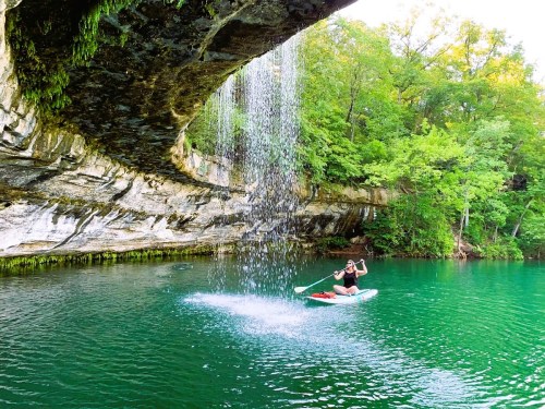 A person kayaking under a waterfall surrounded by lush green trees and clear turquoise water.