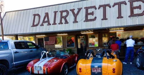 Two classic sports cars parked outside a retro diner named "Dairyette," with people inside and an American flag visible.