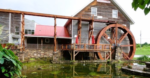 A rustic watermill with a large wooden wheel, surrounded by greenery and a pond, under a cloudy sky.
