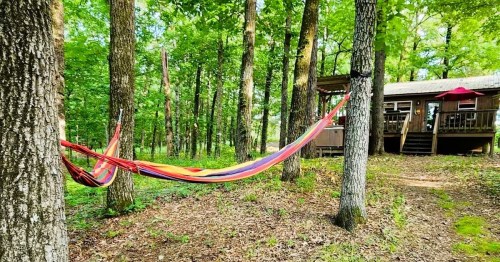 A colorful hammock strung between two trees in a wooded area, with a cabin visible in the background.