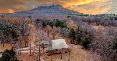 Aerial view of a cabin surrounded by autumn trees, with a mountain in the background under a dramatic sky.