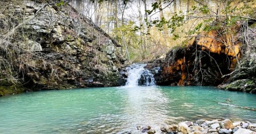 A serene pool of turquoise water surrounded by rocky cliffs and a small waterfall, framed by trees and foliage.