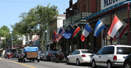 A street lined with shops displaying various flags, including the Confederate flag, with parked cars and trees.