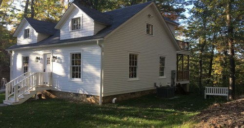 A white two-story house surrounded by trees, featuring a porch and a small bench in the yard.