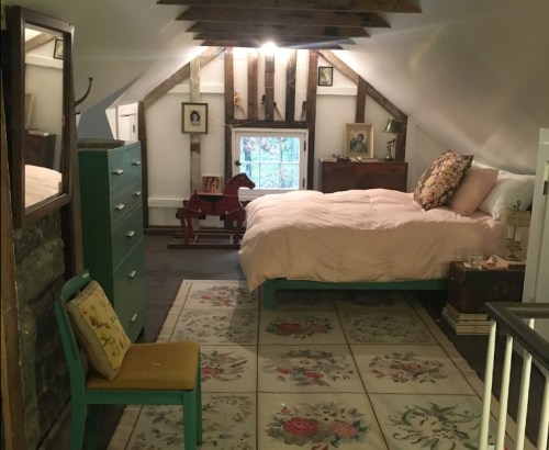 Cozy attic bedroom with a bed, vintage furniture, a rocking horse, and a floral rug under sloped wooden beams.