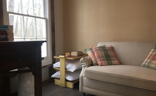 Cozy interior with a beige couch, plaid pillows, a wooden table, and a shelf of books near a window.