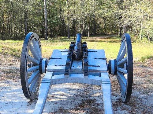 A historic cannon on a wooden carriage, set against a backdrop of trees and grassy terrain.