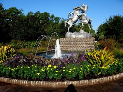 A fountain with a horse statue surrounded by colorful flowers and greenery under a clear blue sky.