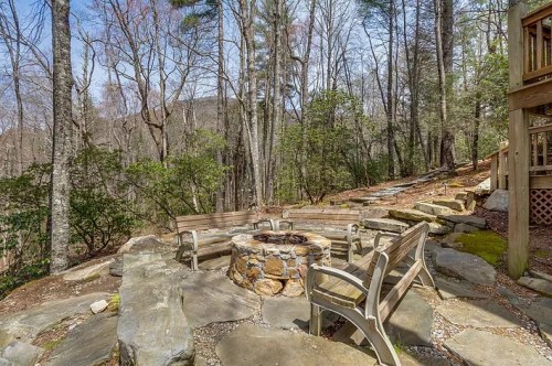 A stone fire pit surrounded by chairs in a wooded area with trees and a clear sky in the background.