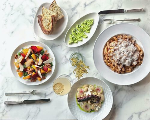 A marble table set with a variety of dishes: pasta, salad, octopus, green sauce, bread, and a drink.