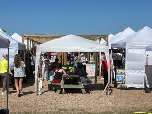 A bustling outdoor market with white tents, people socializing, and a clear blue sky overhead.