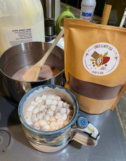 A cozy scene featuring a mug of hot chocolate topped with marshmallows, a pot of chocolate mix, and a bag of cocoa.