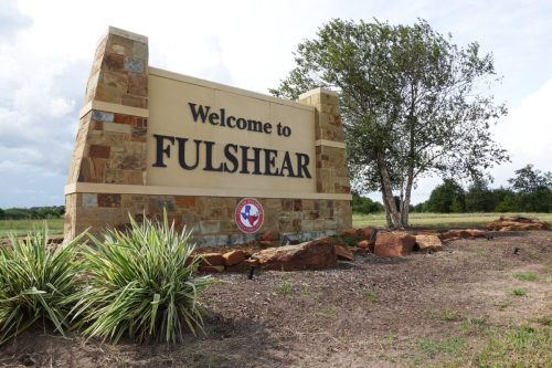 Welcome sign for Fulshear, Texas, featuring stone structure and landscaping under a cloudy sky.