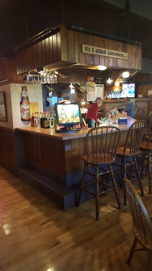 A cozy bar with wooden decor, a drink display, and a sign reading "It's 5 o'clock somewhere."