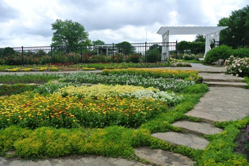 A vibrant garden with colorful flowers and a stone pathway, featuring a white gazebo in the background under cloudy skies.