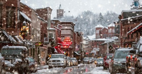 Snowy street scene with shops and cars, festive lights, and falling snowflakes creating a cozy winter atmosphere.