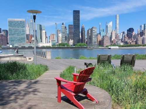 A red Adirondack chair overlooks a river with a city skyline in the background, surrounded by greenery.