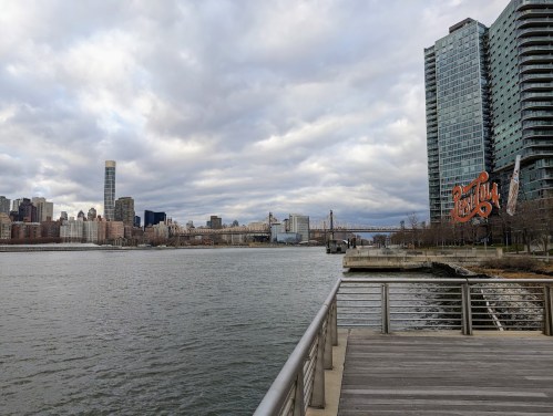 View of a river with a city skyline in the background, featuring a bridge and modern buildings under a cloudy sky.