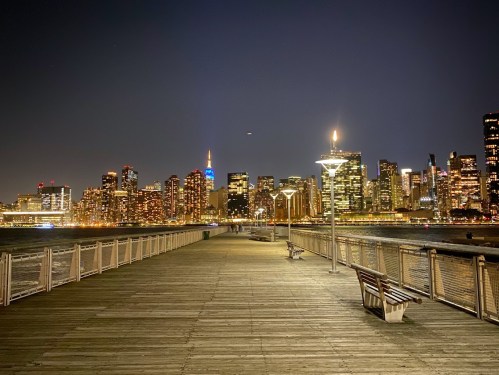 A nighttime view of a city skyline with illuminated buildings, seen from a wooden pier with benches.