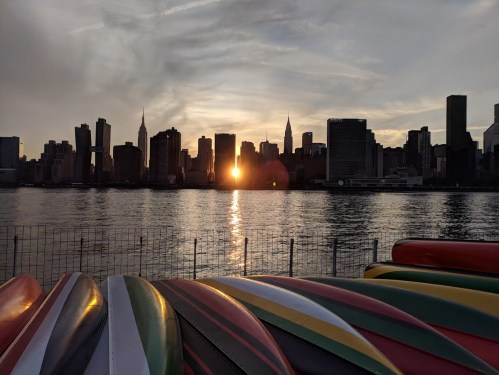 Sunset over a city skyline with the Empire State Building, viewed from a waterfront with colorful kayaks in the foreground.