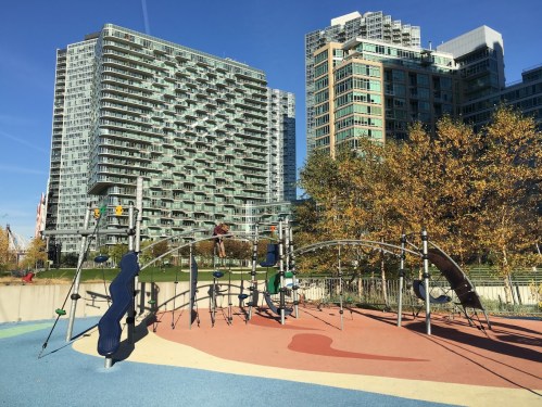 A colorful playground with modern equipment, surrounded by tall buildings and autumn trees under a clear blue sky.
