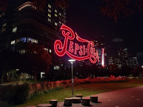 Neon Pepsi-Cola sign illuminated at night, surrounded by a park and city skyline in the background.