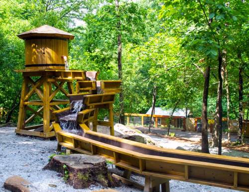 A wooden play structure with a water feature, surrounded by trees and gravel, in a natural outdoor setting.