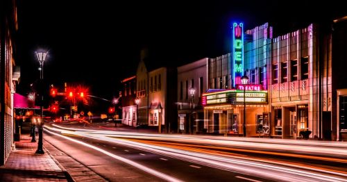 A vibrant theater marquee illuminated at night, with colorful lights and light trails from passing cars on the street.