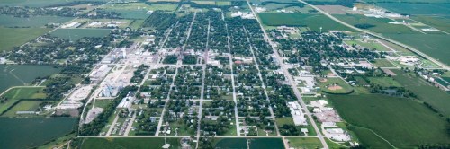 Aerial view of a small town surrounded by green fields and farmland, featuring a grid layout of streets and buildings.