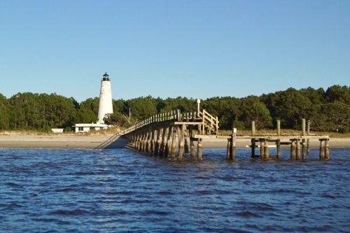 A lighthouse stands in the background, with a wooden pier extending over calm waters and trees lining the shore.