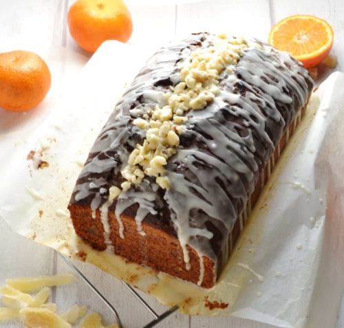 A glazed loaf cake topped with chopped nuts, surrounded by orange slices on a white wooden surface.
