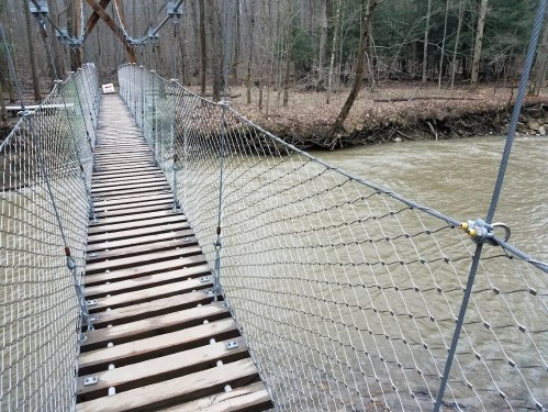 A wooden suspension bridge over a river, surrounded by trees in a forested area.