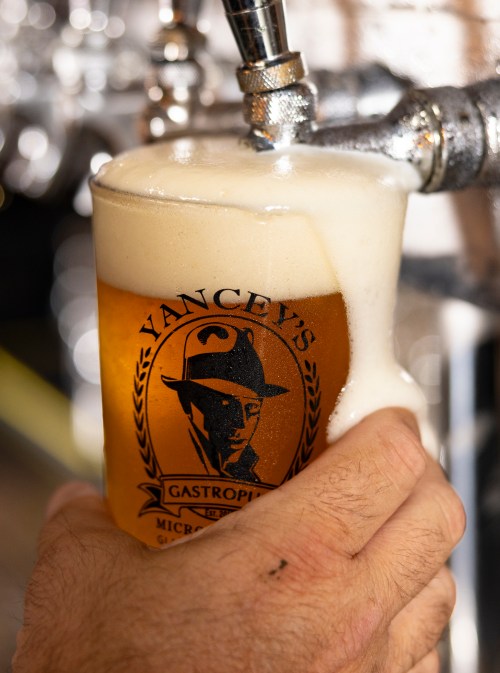 A hand holds a frothy beer in a glass with a logo, as it’s being poured from a tap.