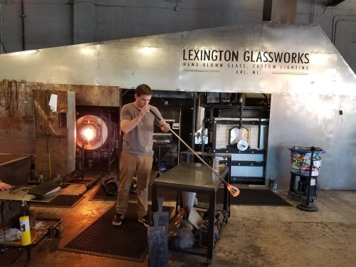 A glassblower shapes molten glass at Lexington Glassworks, with a furnace and tools in the background.