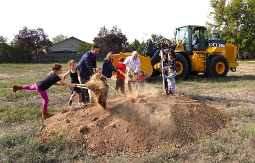 A group of children and adults shovel dirt at a construction site, with a large yellow bulldozer in the background.