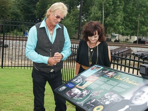 Two people examine a historical plaque outdoors, with a train track visible in the background.