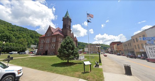 Historic building with a clock tower, surrounded by trees and a street with shops, under a blue sky with clouds.