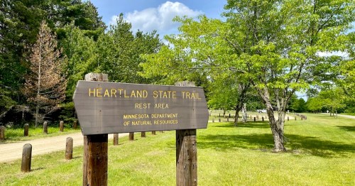 A wooden sign for a Heartland State Trail Rest Area, managed by the Minnesota Department of Natural Resources, stands near a gravel path and green lawn.