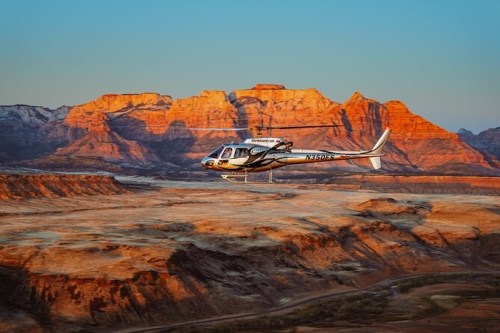 A helicopter flies over rugged, colorful mountains at sunset, casting warm light on the landscape below.