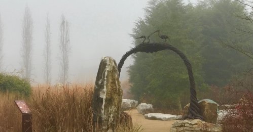 A foggy landscape featuring a stone arch with two birds perched on top, surrounded by tall grasses and trees.