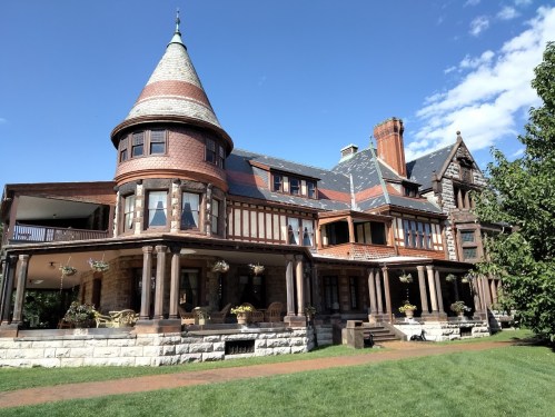 A large, historic mansion with a turret, intricate architecture, and a lush green lawn under a clear blue sky.