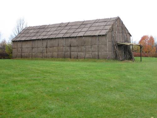 A large, rustic barn made of wooden beams and thatched roof, set on a grassy field with trees in the background.