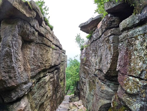 A narrow rocky passageway between towering stone walls, surrounded by greenery and misty atmosphere.