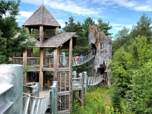 A wooden play structure with a tower and a rock-like formation, surrounded by trees and greenery under a blue sky.