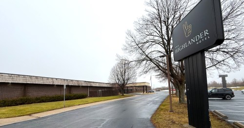 The Highlander Hotel outdoor signage stands next to the driveway and parking lot in front of the Iowa City hotel's brick exterior.