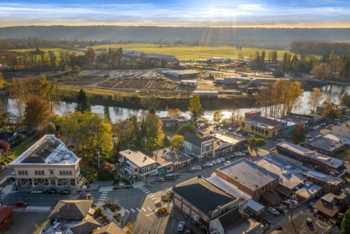 Aerial view of a small town with shops, trees, and a river, surrounded by fields and hills under a sunny sky.