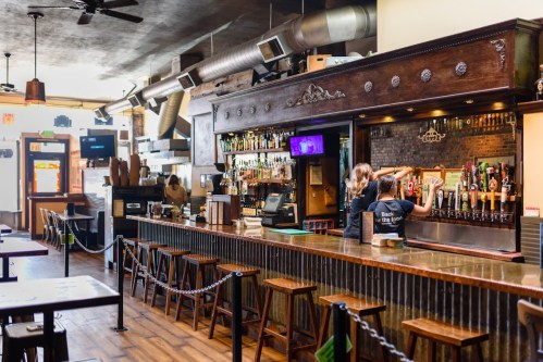 A bar interior with wooden stools, a long counter, and a bartender serving drinks amidst shelves of liquor.