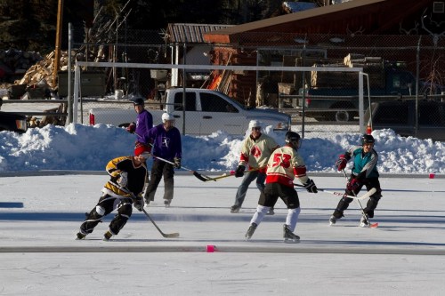 A group of ice hockey players in colorful jerseys skating and playing on an outdoor rink surrounded by snow.